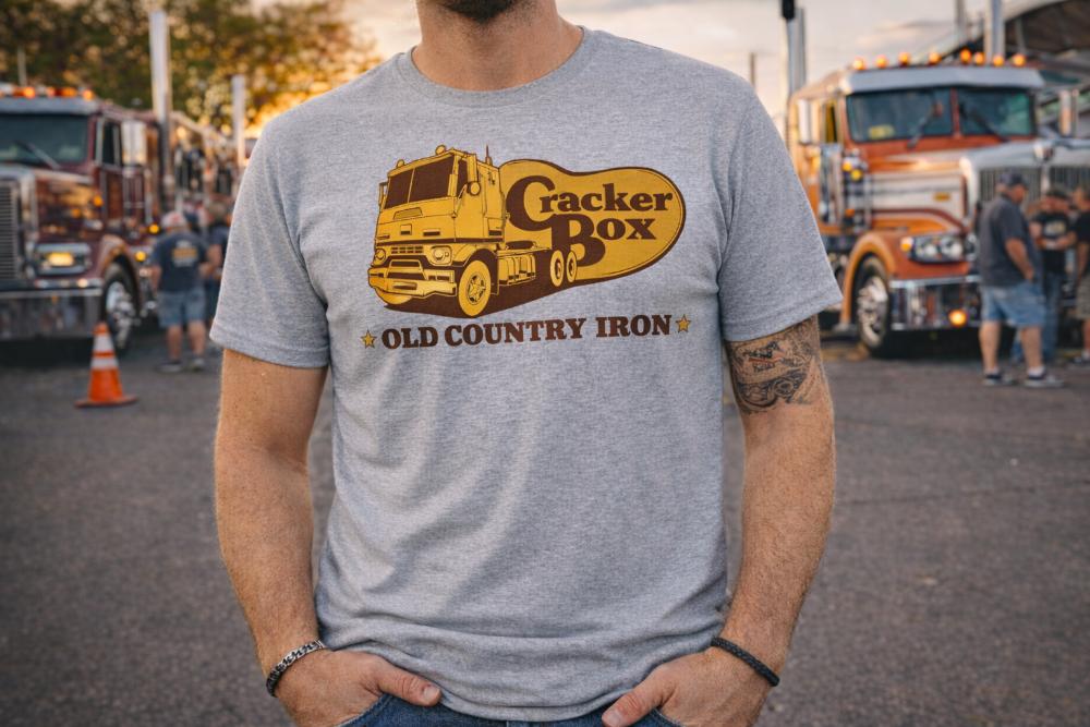 Person wearing a t-shirt with a Cracker Box graphic in an outdoor setting with trucks in the background.