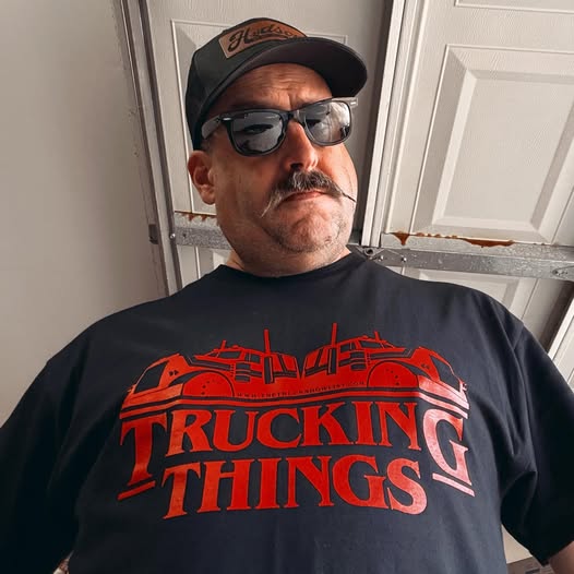 Man wearing a black t-shirt with 'Trucking Things' in red text, sunglasses, and a cap indoors.