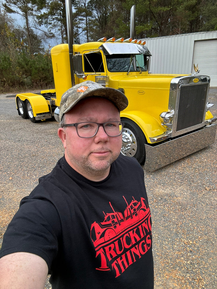 Man wearing a black t-shirt Trucking Things tshirt standing in front of a yellow truck.